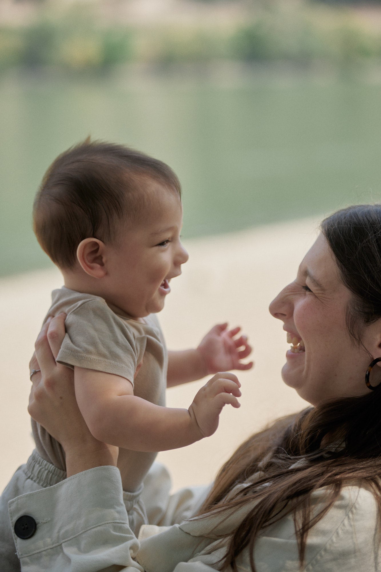 woman holds a baby, both mom and child smiling, outside by the water