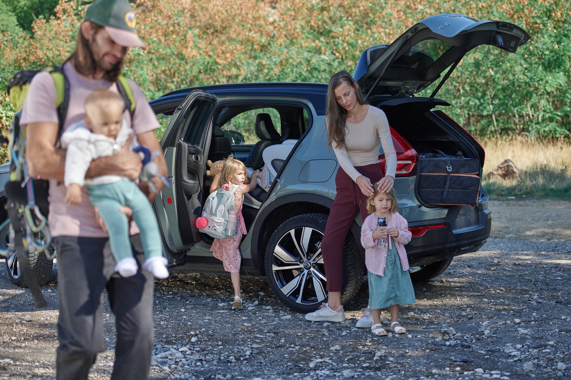 family outdoors in nature, 4-door open, trunk open. Mom does girl's hair while other girl exits car, Dad holds a baby