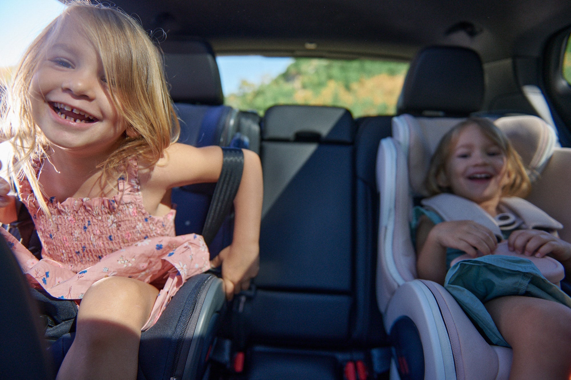 happy girl toddler leans forward in parked car, sibling smiling from safely buckled forward facing babyark car seat