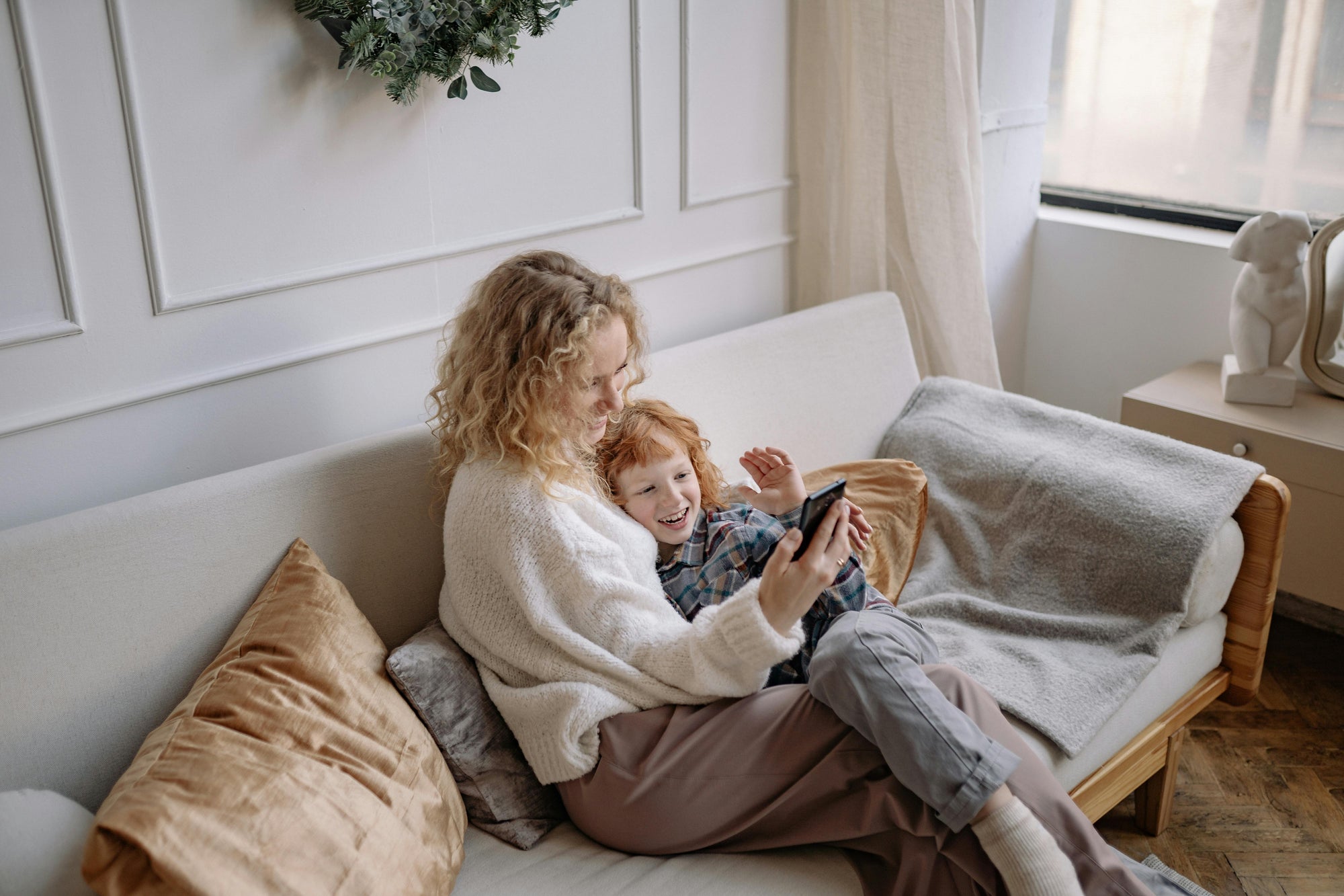 Mom with blonde wavy hair sits on couch with red-headed child on a video call