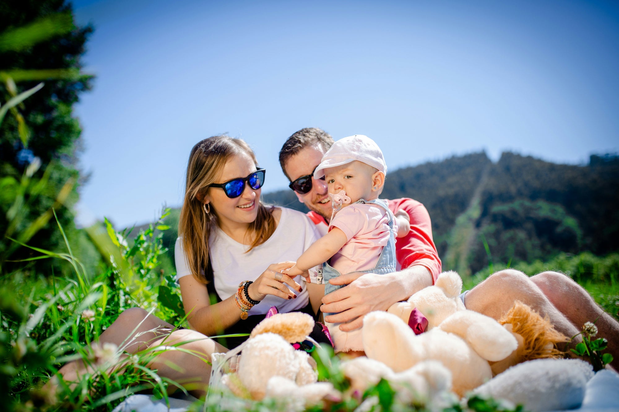 woman and man in sunglasses sit with a cute child in a field surrounded by stuffed animals